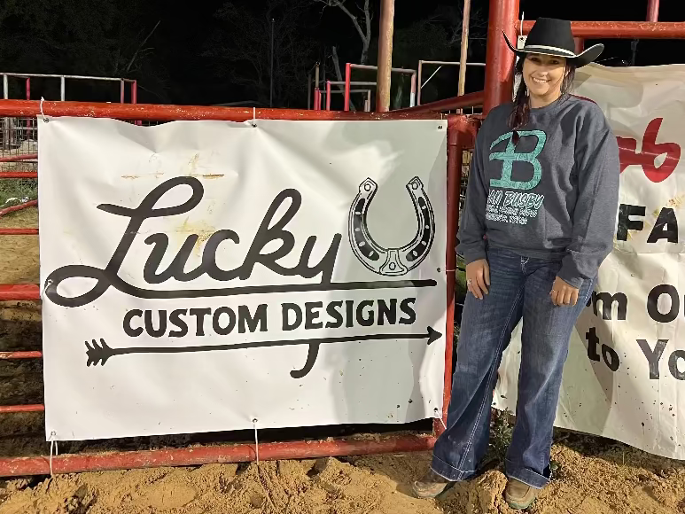 Person wearing a cowboy hat and casual outfit standing next to a 'Lucky Custom Designs' banner at an outdoor event.